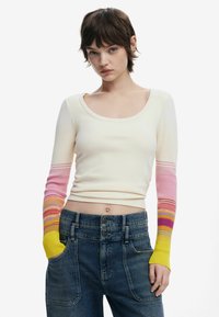 Cremfarbener Pullover mit langem Arm und gerippter Textur. Die Ärmel verlaufen von Pink zu Gelb und haben horizontale Streifen. Kombiniert mit hochtaillierter Jeans.