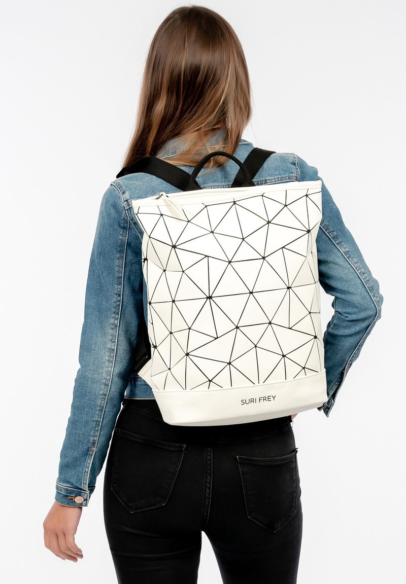 Femme portant un sac à dos blanc à motif géométrique avec des bretelles noires, une veste en jean et un pantalon noir, dos à la caméra.