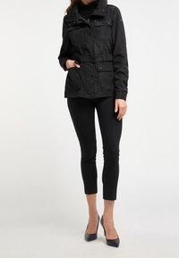 Veste noire légère avec une taille ceinturée, plusieurs poches, des détails à boutons et un col montant, portée avec un pantalon noir ajusté et des talons.