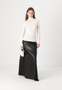 Jupe longue en cuir noir associée à un pull col roulé blanc ajusté. Le mannequin tient un petit sac à main beige, mettant en valeur un design épuré et minimaliste.