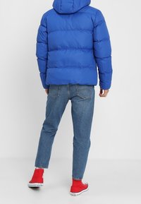 Blauwe gewatteerde jas met een capuchon, met een boxy ontwerp. Draag het over spijkerbroeken en rode high-top sneakers. Zichtbare stiksels en textuur.