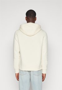 Huppari vaaleanruskeassa värissä, jossa on tilava huppu, ribatut hihansuut ja helma sekä pehmeä tekstuuri. Takana ei ole näkyvää logoa tai suunnitteluviivoja.