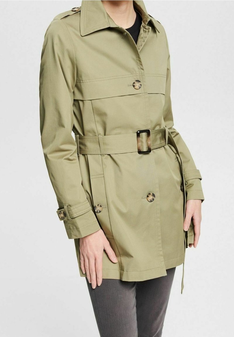 Oliwkowy płaszcz trench wykonany z lekkiego materiału, z paskiem w talii, guzikami i usztywnionym kołnierzem.