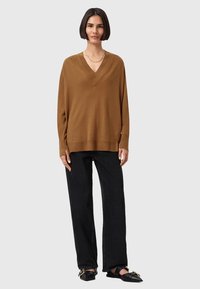 Maglione marrone a V con maniche lunghe, polsini e orlo a costine, abbinato a jeans neri a gamba larga e scarpe nere senza lacci.