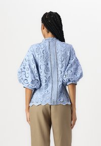 Lyseblå bluse i blonde med pufferm, med blomstermønster, bølget kant og en sentral glidelås på baksiden. Kombinert med beige bukser.