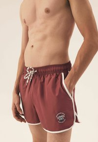 Bordowe szorty kąpielowe z białymi akcentami, elastycznym pasem i sznurkiem do regulacji. Na lewej udzie znajduje się logo "Letnie Wibracje".