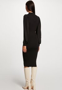 Robe noire côtelée avec de longues manches bouffantes, coupe ajustée et longueur au genou. Assortie à des bottes beiges montantes. Texture lisse et coupe sur mesure.