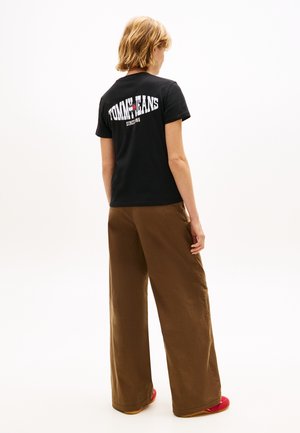 Άτομο με κοντά ξανθά μαλλιά που φοράει μαύρο μπλουζάκι Tommy Jeans, καφέ παντελόνι με φαρδιά πόδια και κόκκινα παπούτσια, στεκόμενο με πλάτη.