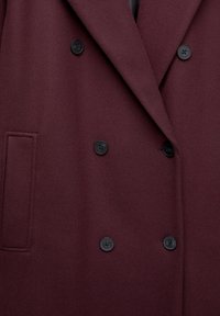 Bordo double-breasted kaput napravljen od teksturiranog materijala, s crnim dugmadi i bočnom džepom za dodatni detalj.