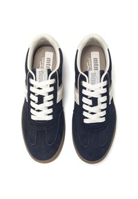 Zapatillas de ante azul marino con cordones blancos y una franja lateral beige. Presentan una parte superior texturizada y una suela de goma marrón. Diseño de lengua plana.