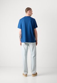Camiseta de algodón azul con manga corta, cuello redondo, combinada con vaqueros azul claro y zapatos beiges. Vista trasera; incluye un tatuaje en el brazo.