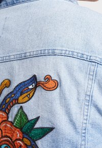 Ljusblå jeansjacka med färgstarkt broderat motiv av en orm, lågor och blad på ryggen.