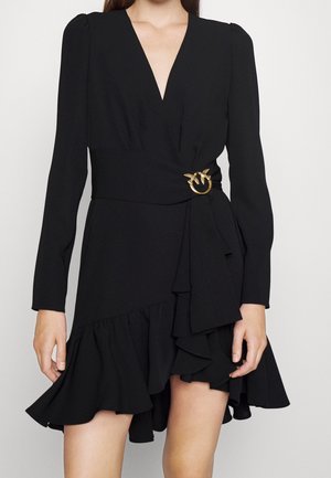 Woman wearing a black V-neck long-sleeve dress with ruffled asymmetrical hem and gold bird buckle detail at the waist.