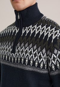 Pulover tricotat bleumarin și verde măslin cu un guler înalt cu nervuri, având un model geometric alb și gri pe piept. Detaliu cu fermoar.