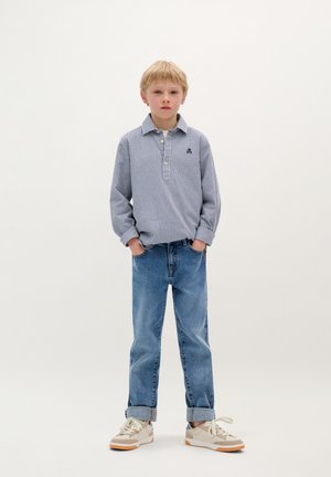 Blauw en wit geruit overhemd met lange mouwen, lichtblauwe jeans en beige sneakers met witte veters; staande pose, handen in de zakken.