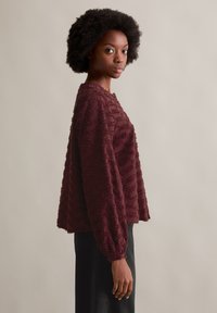 Burgundowa bluzka o fakturowanej powierzchni, z szerokimi rękawami i luźnym krojem, zestawiona z czarnymi spodniami w kontrastowym stylu.