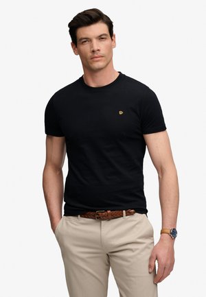 Mand iført sort tætsiddende kortærmet t-shirt, beige bukser med flettet brunt bælte og armbåndsur, stående med hænderne i lommerne.