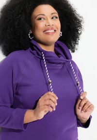 Violetti huppari, jossa on kauluksen yläosa, valkoiset ja violettit nyörit sekä koristeellisia metalliosia. Sileä kangas ja rento leikkaus.