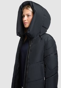 Schwarze Pufferjacke mit einer großen, isolierten Kapuze. Verfügt über Reißverschluss- und Druckknopfverschlüsse, seitliche Taschen und eine gesteppten Textur. Glattes, wasserabweisendes Material.