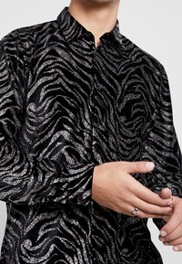 Černá sametová košile s metalickými stříbrnými pruhy ve vzoru zebry, s límečkem na knoflíky a dlouhými rukávy. Hladká textura, přizpůsobený střih.