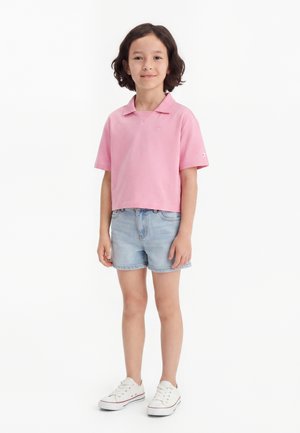 Dítě stojí a usmívá se, má na sobě růžové polo tričko s krátkým rukávem, světle modré džínové kraťasy a bílé tenisky na jednobarevném bílém pozadí.