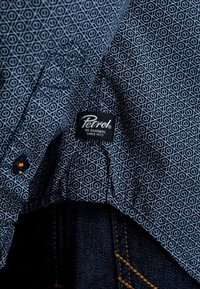 Chemise bleu foncé à motifs avec une étiquette portant la mention "Petrol No Barriers Since 1973", associée à un jean en denim foncé avec des coutures visibles.