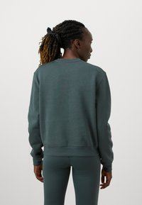 Tmavomodrá mikina so zaobleným výstrihom a rebrovanými manžetami, vyrobená z jemného materiálu. Má voľný strih a je nosená s podpornými legínami.