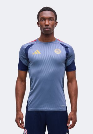 Homme debout face à l'avant portant un maillot de sport bleu et marine du Bayern Munich et un short marine sur un fond uni.