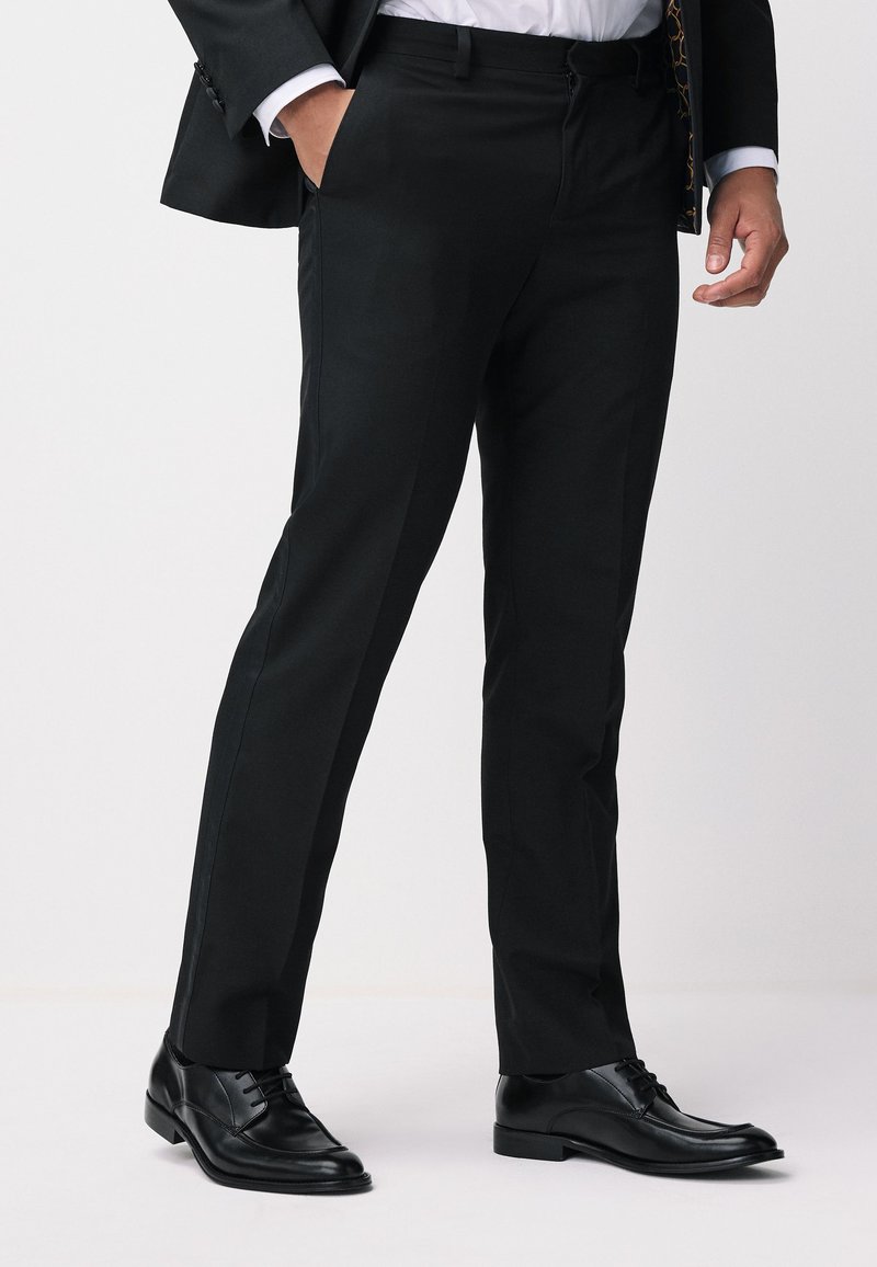 Homme en pantalon de costume noir, chaussures de ville noires et veste sombre, une main dans la poche sur fond blanc.