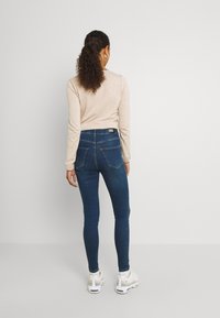 Mujer con cabello rizado, vestida con una blusa de manga larga beige, jeans ajustados azules oscuros y deportivas blancas, de pie frente a una pared clara y lisa.