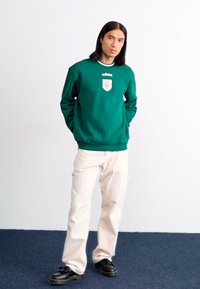 En ung mann står med hendene i lommene, iført en grønn Adidas hettegenser, stripete bukser og svarte sko mot en enkel bakgrunn.