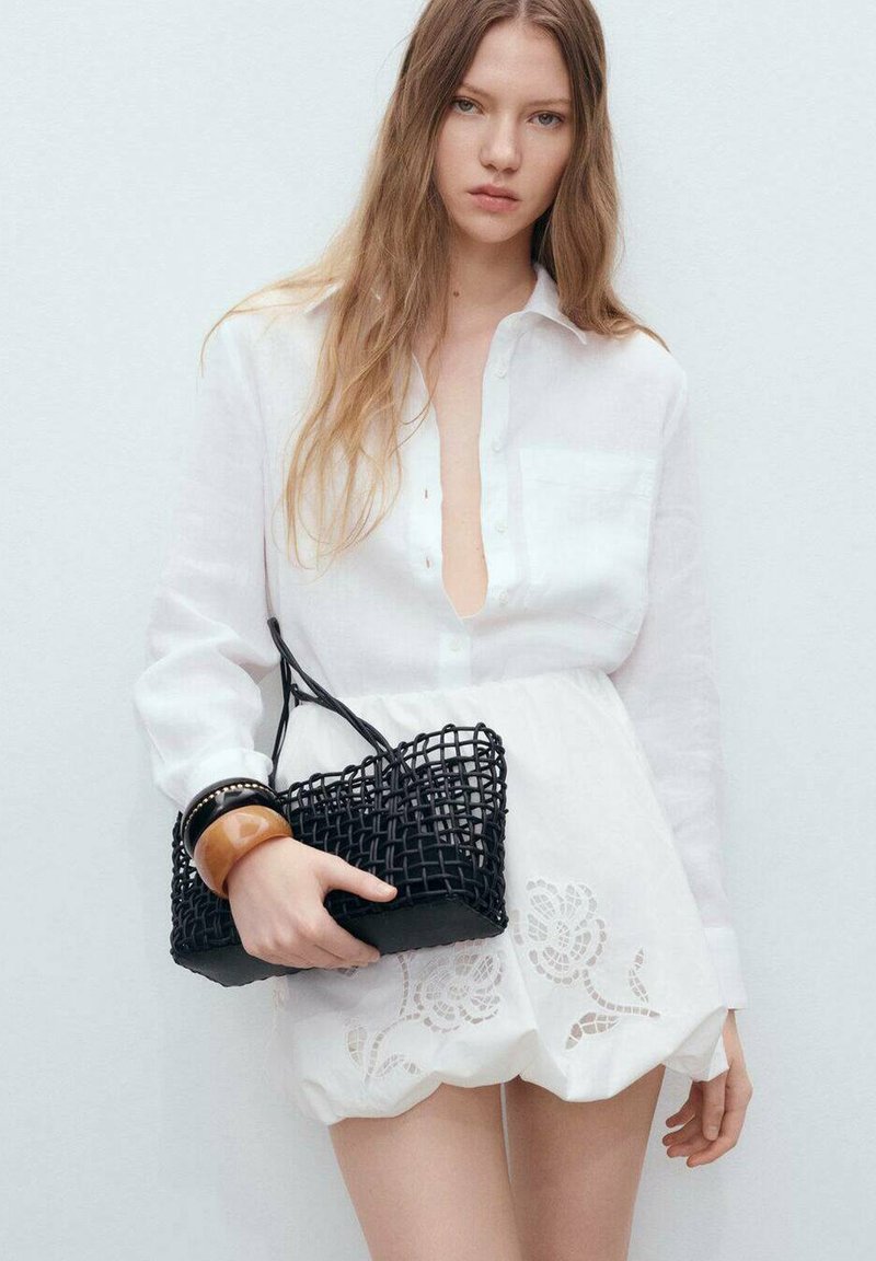 Camicia bianca con scollo a V profondo, indossata sopra una gonna bianca con ricami floreali. Tiene una borsa a tracolla nera intrecciata con dettagli.