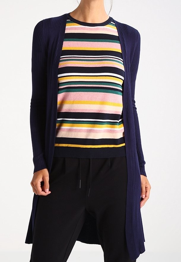 Pitkä laivastonsininen ribbineuletakki monivärisen raidoitetun villapaidan päällä, jossa on vaaleanpunaisia, keltaisia, vihreitä ja sinisiä raitoja, yhdistetty mustiin housuihin.