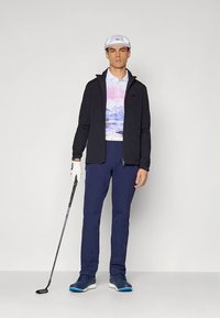 Moška golf obleka, ki vključuje črno jopico z zadrgo, polo majico s pastelno gorskim potiskom, mornarsko modre hlače, belo kacket in golf čevlje z modrimi poudarki.