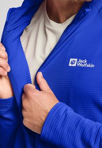 Blaues Zip-Jacket mit gerippter Textur und Logo auf der Brust, kombiniert mit einer hellbeigen Basisschicht darunter, die das Design des Kleidungsstücks präsentiert.