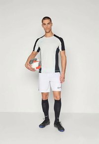 Meeste sportlik riietus, mis sisaldab heledat halli lühikeste varrukatega särki mustade aktsentidega, valgeid lühikesi pükse ja musti sokke, hoides jalgpalli.