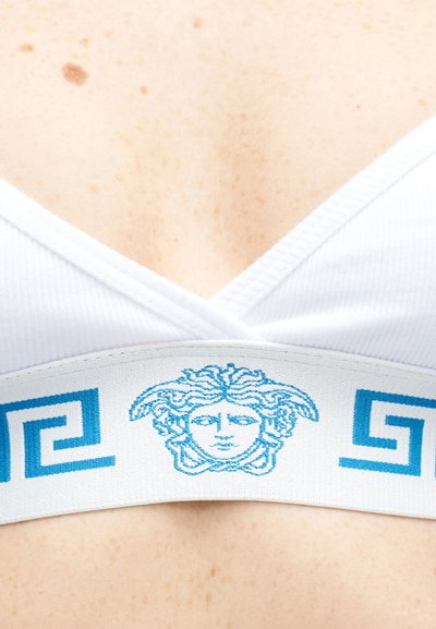 Λευκό αθλητικό σουτιέν με μπλε λογότυπο κεφαλής Μέδουσας και γεωμετρικά σχέδια στη λαστιχένια ταινία, φορεμένο σε δέρμα με φακίδες.