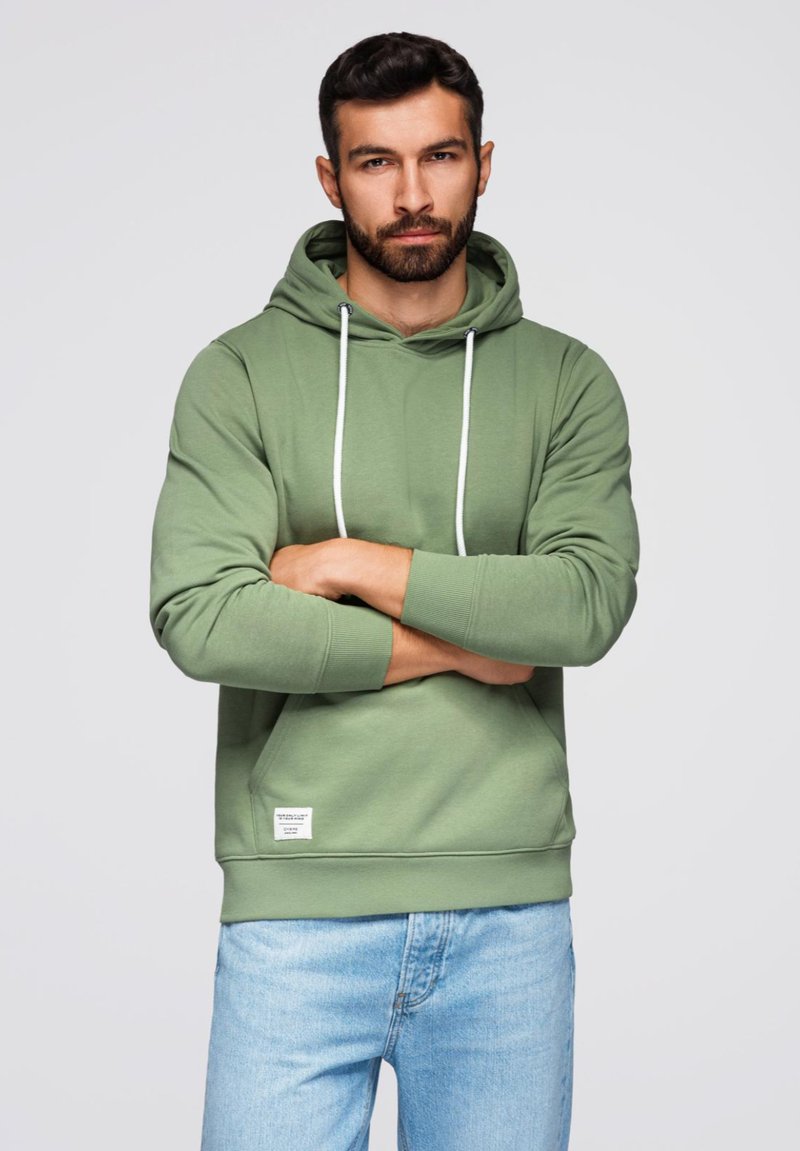 Sudadera de algodón verde con capucha ajustable, bolsillo frontal tipo canguro y puños acanalados. Combinada con jeans de color azul claro, mostrando un corte relajado.