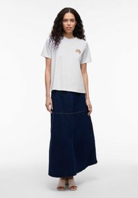 Lichtgrijze T-shirt met een kleine grafische print, gecombineerd met een donkere denim maxi rok met een horizontale naad en een relaxte pasvorm. Gesloten beige sandalen.