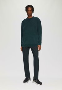 Teal getextureerde sweatshirt met een ronde hals, gecombineerd met donkere broek en zwarte sneakers met witte zolen, weergegeven tegen een lichte achtergrond.