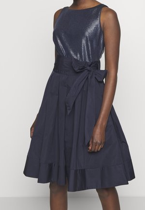 Mouwloze marineblauwe jurk met glanzend aansluitend lijfje, brede tailleband gestrikt in een strik, en geplooide A-lijn rok die boven de knie valt.