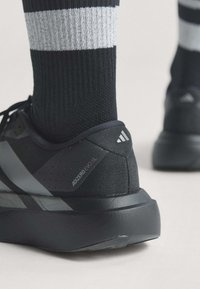Κοντινή λήψη του μαύρου αθλητικού παπουτσιού Adidas Adizero Evo SL, φορεμένου με μαύρη και άσπρη ριγέ κάλτσα σε ανοιχτό φόντο.