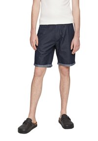 Shorts en denim bleu foncé avec des ourlets roulés, présentant une coupe décontractée et une texture lisse, associés à des sabots noirs à enfiler.