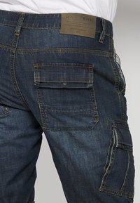 Pantalon cargo en jean bleu foncé avec plusieurs poches à rabat et un patch de marque en cuir marron sur la ceinture arrière.