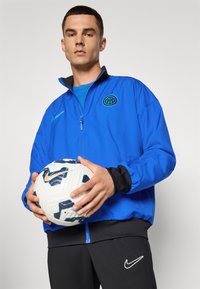 Blå zip-up jakke med sorte accenter og logo, parret med en blå skjorte og sorte bukser. Holder en hvid fodbold med blå og sorte mønstre.