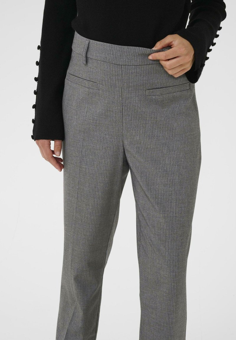 Personne portant un pantalon gris à fines rayures et un pull noir, ajustant la taille d'une main, debout devant un fond uni.