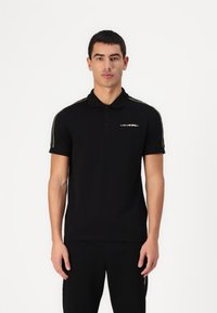 Svart pikétröja med krage, försedda med gyllene sidskivor och en logotyp på bröstet. Tillverkad av ett mjukt, stretchigt material.