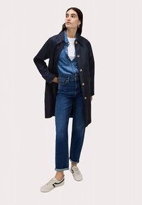 Marineblauwe regenjas met grote knopen, gedragen over een denim blouse en wit T-shirt. Gecombineerd met rechte jeans en grijze sneakers met strepen.
