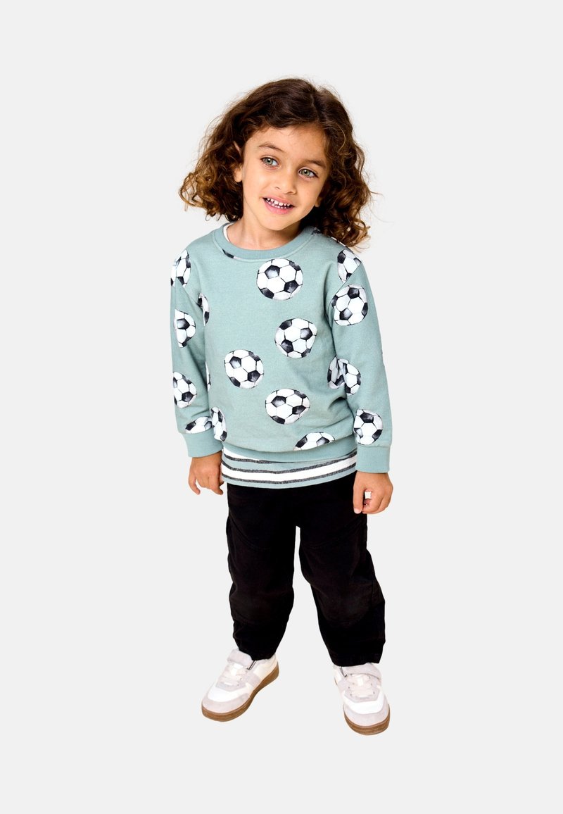 Jeune enfant aux cheveux bouclés portant un sweat-shirt bleu clair avec des imprimés de ballons de football, un pantalon noir et des baskets blanches, debout sur un fond uni.