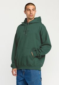 Sudadera verde, material de algodón, capucha con cordón ajustable, bolsillo frontal, logo negro "VOLCOM" en el pecho, puños acanalados, corte casual, combinada con jeans azules.
